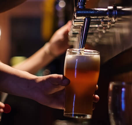 Hand holding a glass almost filled with beer while filling it all the way up with beer from a tap inside a bar